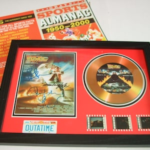 May include: A framed gold record with a black and white image of a car and flames. The record is titled "Back to the Future" and has a red background. The frame also includes a black and white movie poster with the title "Back to the Future" and several signatures. The poster is surrounded by a red mat and a black frame. The frame also includes three small black and white film strips. The frame has a black license plate with the text "JUL CALIFORNIA OUTATIME".