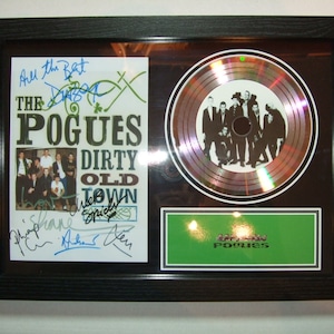 Puede incluir: Un disco de platino enmarcado para el álbum "Dirty Old Town" de The Pogues. El disco está rodeado por un marco negro y una moldura blanca. La portada del álbum es visible en el marco, junto con las firmas de los miembros de la banda. El disco es un disco plateado con un efecto holográfico.