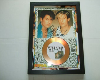 May include: A framed gold record award featuring a photograph of two men in the top half of the frame. The record is a gold disc with the text "WHAM! The Video" printed on it. The men's signatures are visible in the photograph.