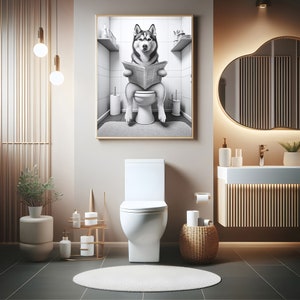 May include: A black and white photo of a husky dog sitting on a toilet and reading a newspaper. The photo is framed and hanging on a wall in a bathroom with a white toilet, a brown wicker basket, and a white sink.