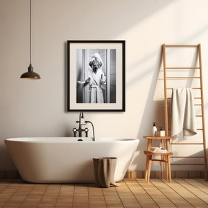 May include: A black and white photograph of a dog wearing a white bathrobe and a towel on its head, standing in a bathroom doorway. The photo is framed in a black frame and hanging on a white wall.