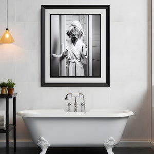May include: A black and white photograph of a dog wearing a white bathrobe and a towel on its head, holding a bottle of soap. The photo is framed in a black frame and hanging on a white tiled wall above a white clawfoot bathtub.