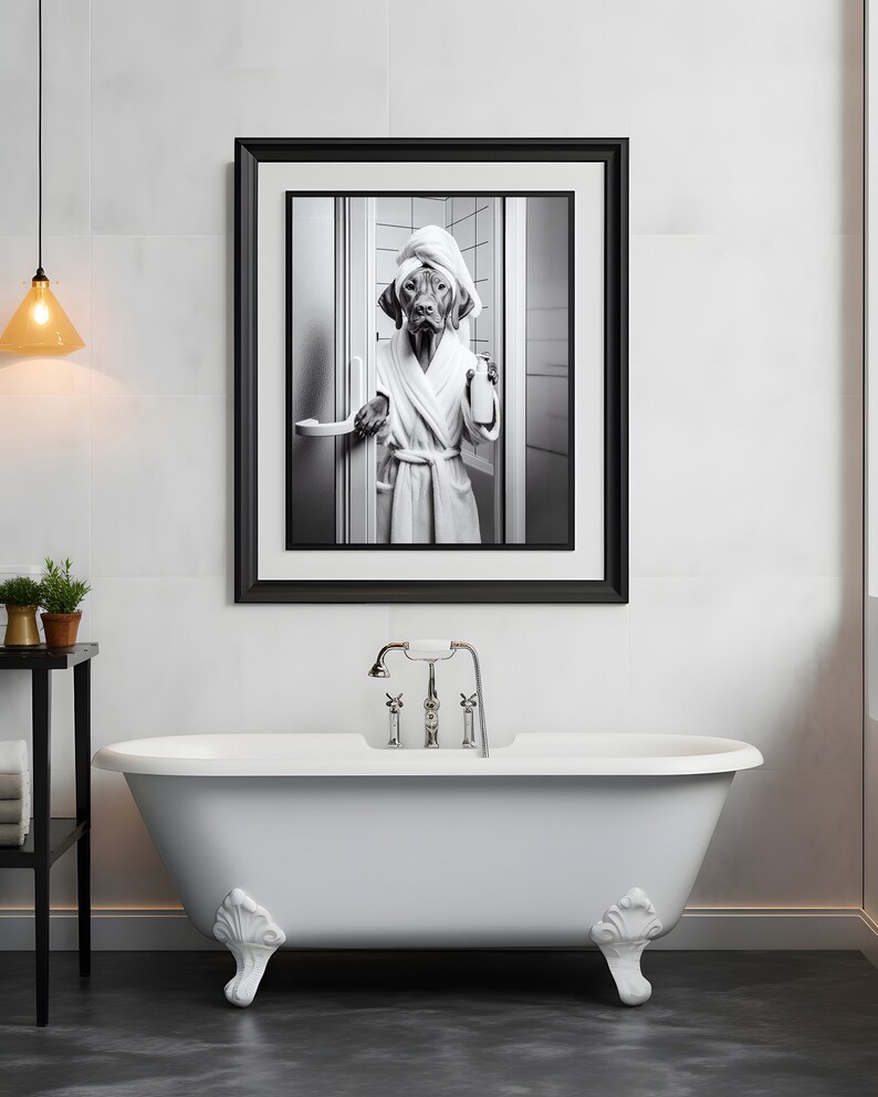 May include: A black and white photograph of a dog wearing a white bathrobe and a towel on its head, holding a bottle of soap. The photo is framed in a black frame and hanging on a white tiled wall above a white clawfoot bathtub.