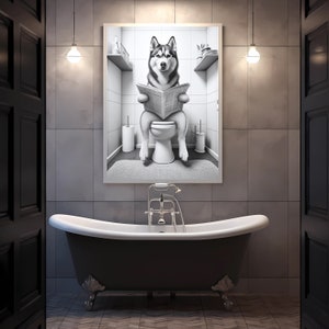 May include: A black and white photo of a husky dog sitting on a toilet and reading a newspaper. The photo is framed and hanging on a bathroom wall above a black bathtub.