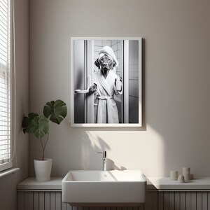 May include: A black and white photograph of a dog wearing a white bathrobe and a towel on its head, standing in a bathroom doorway.