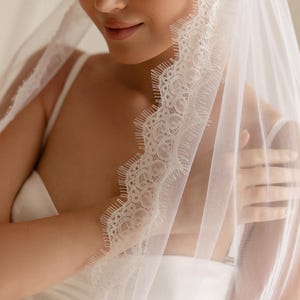 May include: A sheer white bridal veil featuring detailed lace edging. The veil is designed to be worn over the head, with the lace adding a decorative element. A classic wedding accessory, the veil is a symbol of tradition and beauty.