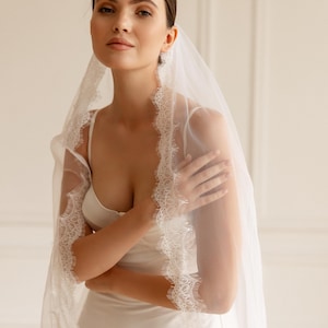Puede incluir: Una novia con un vestido de novia blanco y un velo transparente con delicados bordes de encaje. El velo cae sobre los hombros, creando un look elegante y rom&aacute;ntico. El vestido tiene un dise&ntilde;o sencillo y fluido.