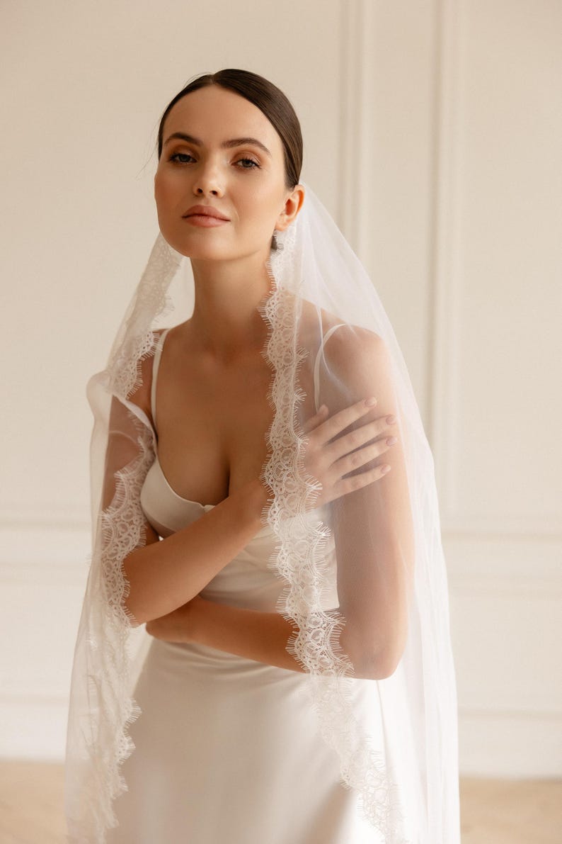 Puede incluir: Una novia con un vestido de novia blanco y un velo transparente con delicados bordes de encaje. El velo cae sobre los hombros, creando un look elegante y rom&aacute;ntico. El vestido tiene un dise&ntilde;o sencillo y fluido.