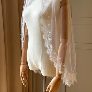 Puede incluir: Un velo de novia blanco transparente con delicados bordes de encaje. El velo est&aacute; drapeado sobre un maniqu&iacute;, mostrando su dise&ntilde;o fluido y sus intrincados detalles. El borde de encaje a&ntilde;ade un toque de elegancia al accesorio nupcial.
