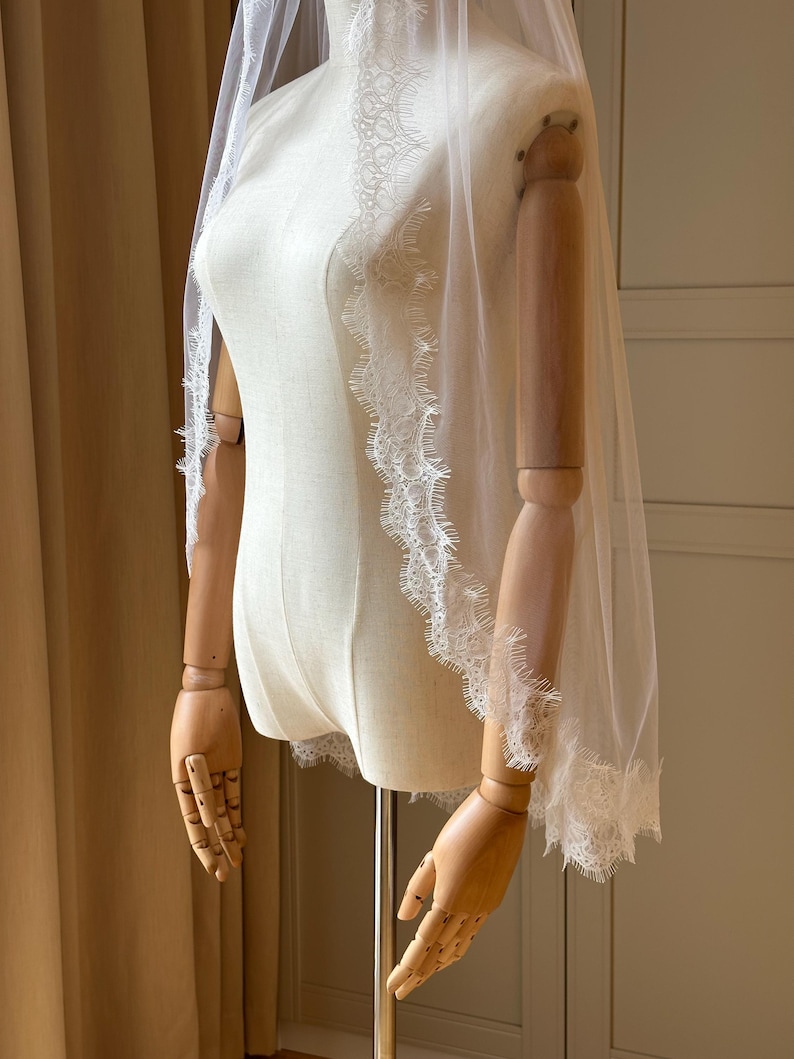 Puede incluir: Un velo de novia blanco transparente con delicados bordes de encaje. El velo est&aacute; drapeado sobre un maniqu&iacute;, mostrando su dise&ntilde;o fluido y sus intrincados detalles. El borde de encaje a&ntilde;ade un toque de elegancia al accesorio nupcial.