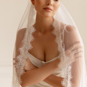 Puede incluir: Un velo de novia blanco con delicados bordes de encaje. El velo est&aacute; drapeado sobre un vestido blanco. Los brazos de la novia est&aacute;n cruzados. El fondo es de un color suave y neutro.