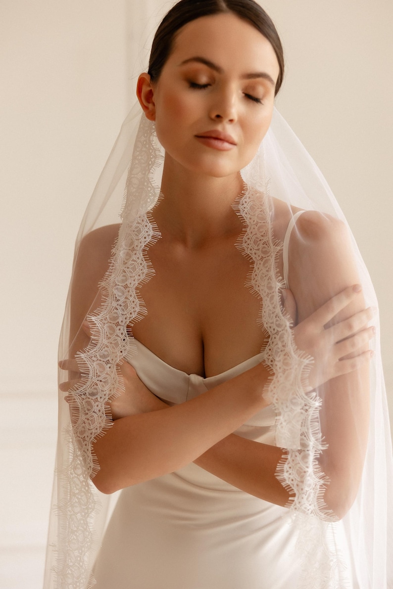 Puede incluir: Un velo de novia blanco con delicados bordes de encaje. El velo est&aacute; drapeado sobre un vestido blanco. Los brazos de la novia est&aacute;n cruzados. El fondo es de un color suave y neutro.