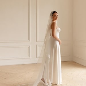 May include: A white wedding dress with a long, flowing skirt and a sweetheart neckline. The bride is wearing a long, sheer veil with delicate lace trim. The dress is simple and elegant, perfect for a classic wedding.