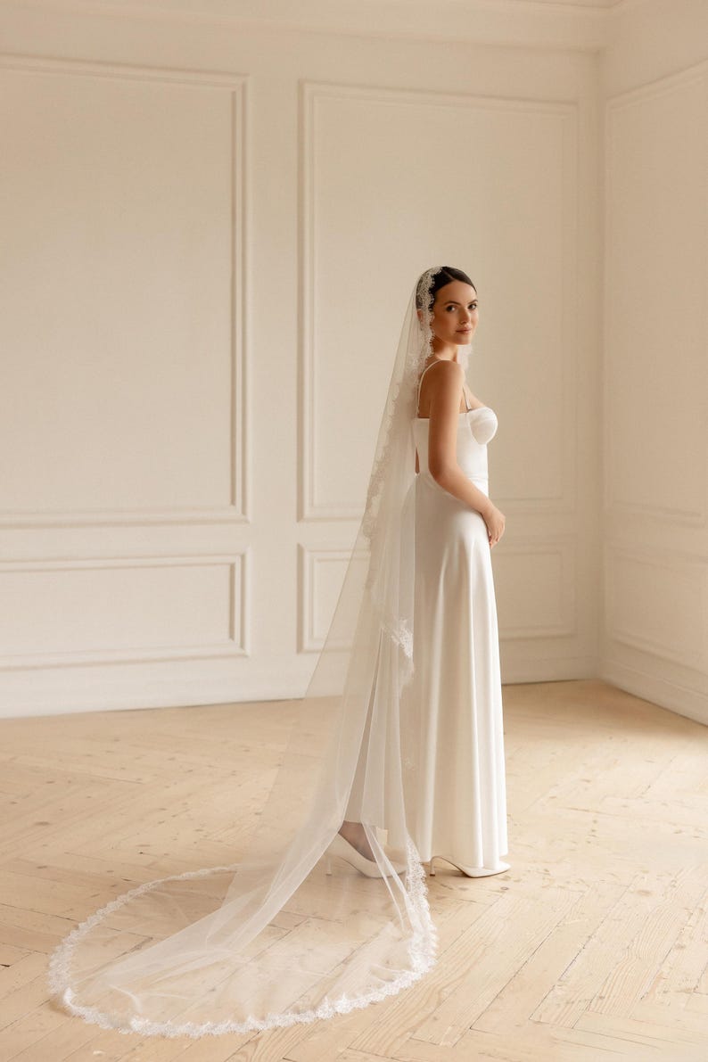 May include: A white wedding dress with a long, flowing skirt and a sweetheart neckline. The bride is wearing a long, sheer veil with delicate lace trim. The dress is simple and elegant, perfect for a classic wedding.