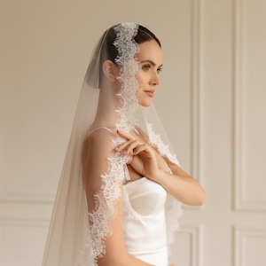 May include: A sheer white bridal veil with delicate lace trim. The veil is draped over the head and shoulders, creating a soft, elegant look. The bride is wearing a white satin dress.