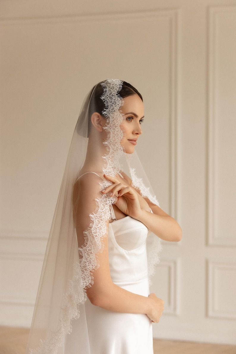 May include: A sheer white bridal veil with delicate lace trim. The veil is draped over the head and shoulders, creating a soft, elegant look. The bride is wearing a white satin dress.