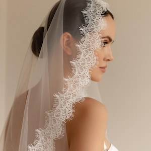May include: A sheer white bridal veil with a delicate, scalloped lace trim. The veil is draped over the head and shoulders, showcasing the intricate lace design. The bride is wearing a white dress.
