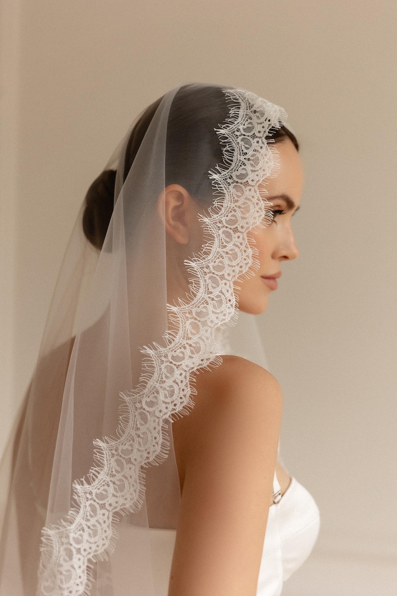 May include: A sheer white bridal veil with a delicate, scalloped lace trim. The veil is draped over the head and shoulders, showcasing the intricate lace design. The bride is wearing a white dress.