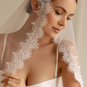 May include: A white bridal veil with delicate lace trim. The lace edge features a scalloped design, adding a touch of elegance. The veil is draped over the head and shoulders, creating a soft, romantic look. Perfect for weddings and special occasions.