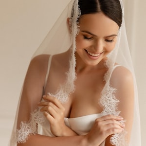 May include: A sheer white bridal veil with delicate lace trim. The veil is draped over the head and shoulders, creating a soft, romantic look. The lace edging adds a touch of elegance to the design. The bride is wearing a white dress.