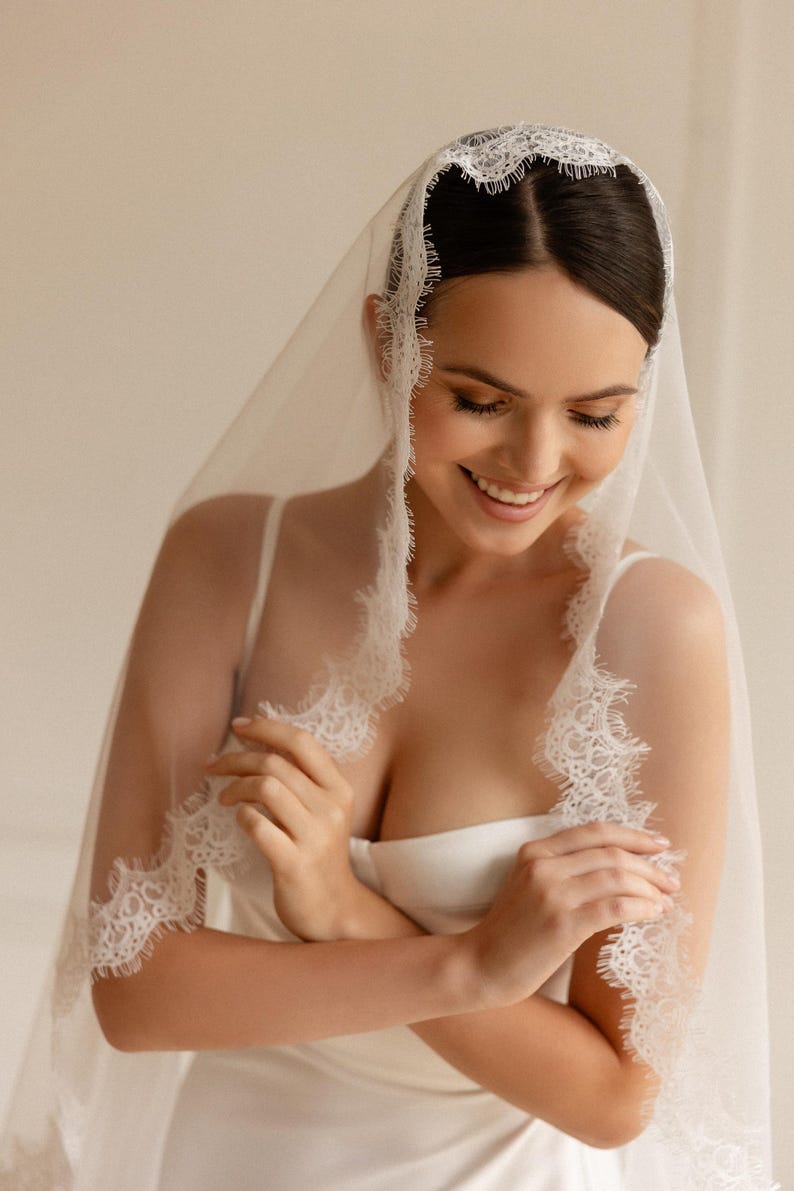 May include: A sheer white bridal veil with delicate lace trim. The veil is draped over the head and shoulders, creating a soft, romantic look. The lace edging adds a touch of elegance to the design. The bride is wearing a white dress.