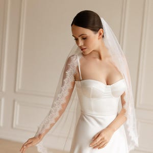 K&ouml;nnte beinhalten: Ein wei&szlig;es Brautkleid mit herzf&ouml;rmigem Ausschnitt und Spaghettitr&auml;gern. Das Kleid wird mit einem transparenten Schleier mit zarter Spitze kombiniert. Die Braut tr&auml;gt das Kleid in einem lichtdurchfluteten Raum.