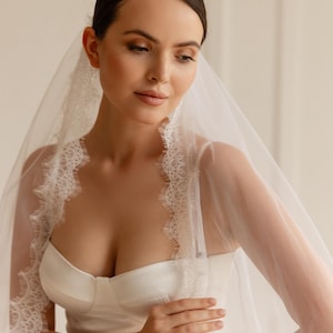 May include: A bride in a white satin wedding dress and a sheer veil with intricate lace edging. The veil drapes around the face and shoulders, creating a soft, romantic effect. The dress features a fitted bodice and a simple, elegant design.