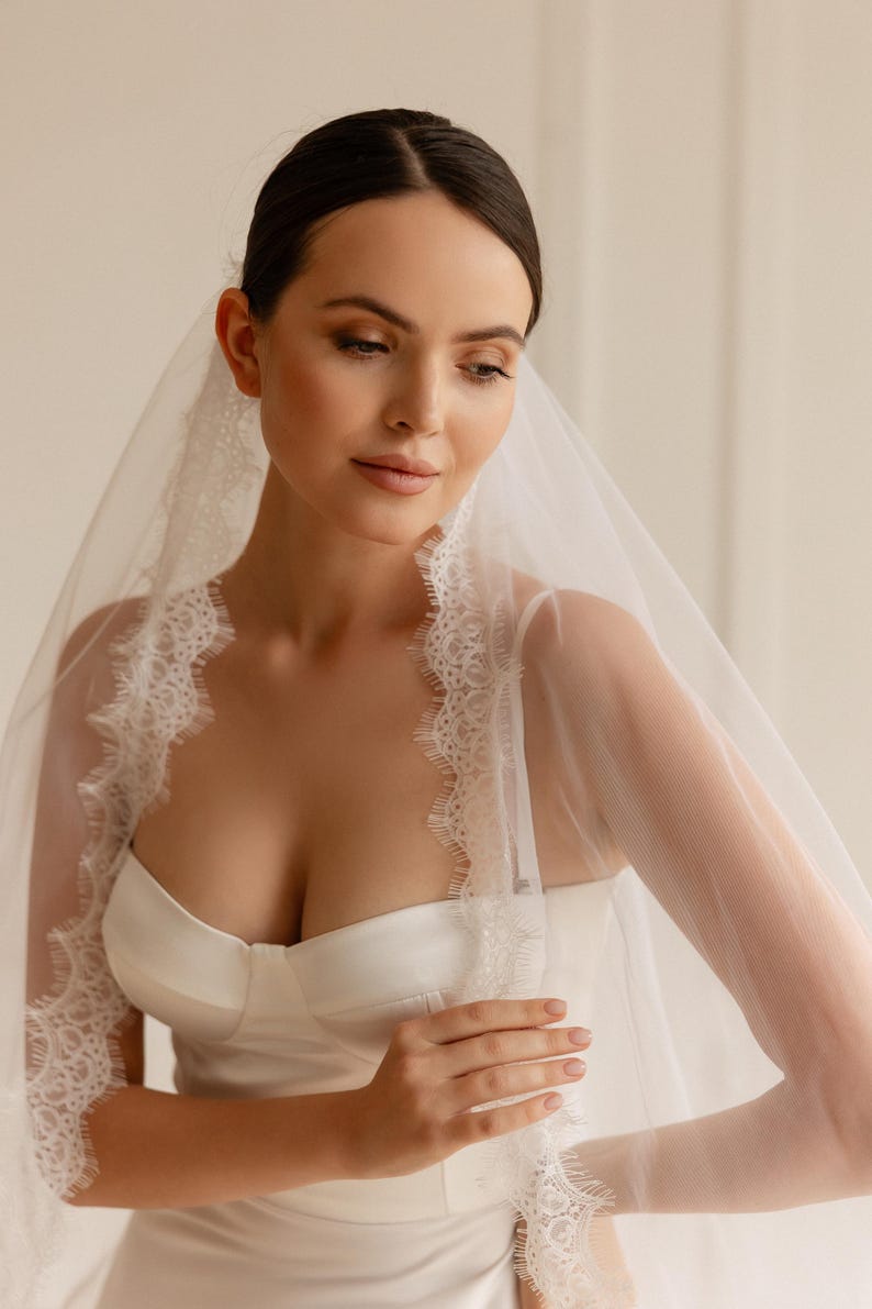 May include: A bride in a white satin wedding dress and a sheer veil with intricate lace edging. The veil drapes around the face and shoulders, creating a soft, romantic effect. The dress features a fitted bodice and a simple, elegant design.