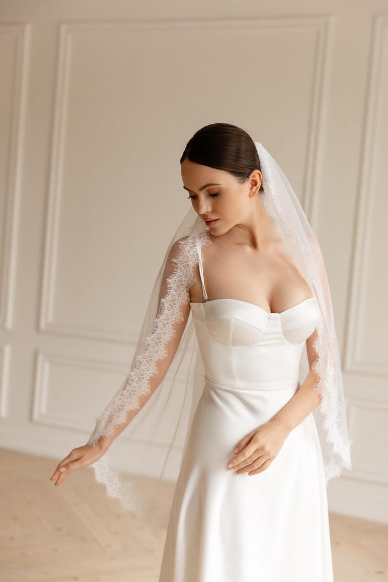 Puede incluir: Vestido de novia de sat&eacute;n blanco con escote coraz&oacute;n y tirantes finos. La novia lleva un velo transparente con delicados bordes de encaje. El velo est&aacute; drapeado sobre los hombros y los brazos. El vestido tiene un corpi&ntilde;o ajustado y una falda fluida.