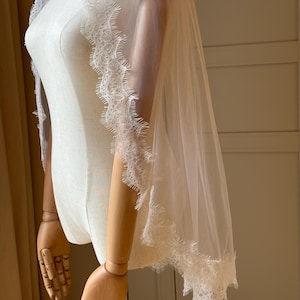Pu&ograve; includere: Un velo da sposa bianco trasparente con delicate finiture in pizzo lungo i bordi. Il velo &egrave; drappeggiato su un manichino, mettendo in mostra l'intricato design in pizzo e il tessuto fluente del velo. La finitura in pizzo aggiunge un tocco di eleganza.