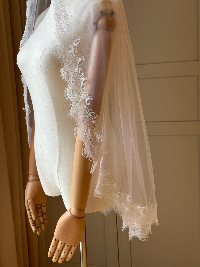 Pu&ograve; includere: Un velo da sposa bianco trasparente con delicate finiture in pizzo lungo i bordi. Il velo &egrave; drappeggiato su un manichino, mettendo in mostra l'intricato design in pizzo e il tessuto fluente del velo. La finitura in pizzo aggiunge un tocco di eleganza.