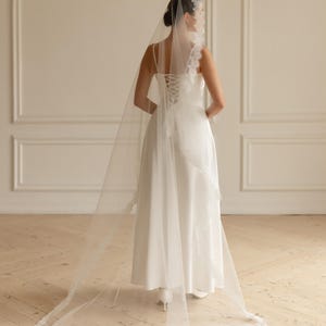 May include: A bride in a white wedding dress with a long, sheer veil. The dress has a corset back and lace detailing. The veil is trimmed with delicate lace and drapes to the floor.