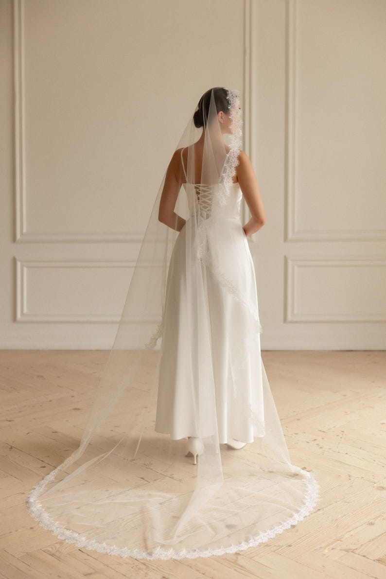 May include: A bride in a white wedding dress with a long, sheer veil. The dress has a corset back and lace detailing. The veil is trimmed with delicate lace and drapes to the floor.