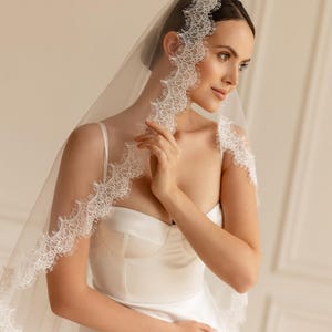 May include: A bride wearing a white wedding dress and veil. The veil is sheer and trimmed with delicate lace. The dress has a sweetheart neckline and a fitted bodice. The bride is looking to the side.