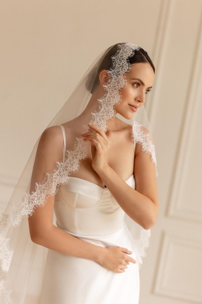May include: A bride wearing a white wedding dress and veil. The veil is sheer and trimmed with delicate lace. The dress has a sweetheart neckline and a fitted bodice. The bride is looking to the side.