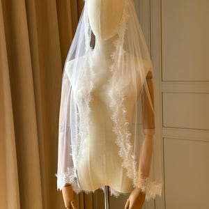 K&ouml;nnte beinhalten: Ein wei&szlig;er Brautschleier mit zarten Spitzenbesatz wird auf einer Schaufensterpuppe pr&auml;sentiert. Der Schleier ist transparent, wodurch die Form der Puppe teilweise sichtbar ist. Der Spitzenbesatz verleiht dem Brautaccessoire Eleganz.