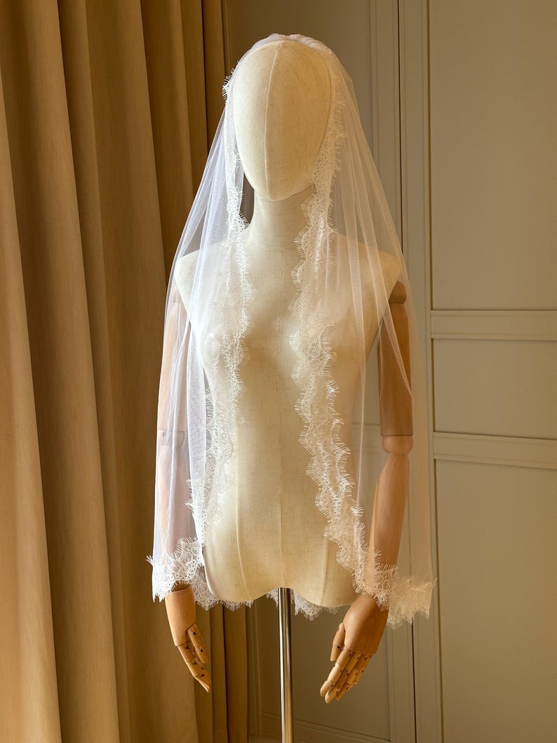 K&ouml;nnte beinhalten: Ein wei&szlig;er Brautschleier mit zarten Spitzenbesatz wird auf einer Schaufensterpuppe pr&auml;sentiert. Der Schleier ist transparent, wodurch die Form der Puppe teilweise sichtbar ist. Der Spitzenbesatz verleiht dem Brautaccessoire Eleganz.