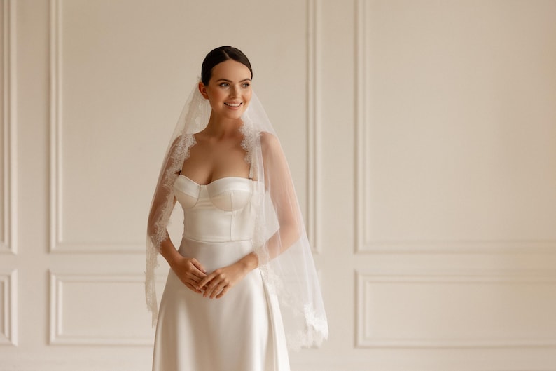 May include: A bride in a white satin wedding dress and veil. The strapless bodice and A-line skirt create an elegant silhouette. The veil is trimmed with delicate lace, adding a touch of romance to the bridal look.
