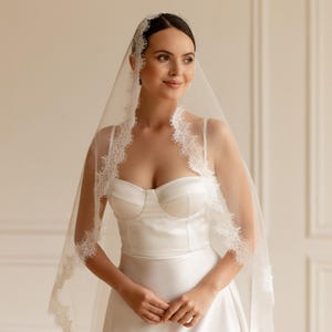 May include: A bride in a white satin wedding dress with a sweetheart neckline and a flowing skirt. She wears a sheer veil with delicate lace trim. The veil frames her face and shoulders, creating an elegant bridal look.