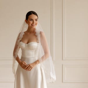 Puede incluir: Una novia con un vestido de novia de sat&eacute;n blanco y velo. El corpi&ntilde;o sin tirantes y la falda en forma de A crean una silueta elegante. El velo est&aacute; adornado con delicado encaje, a&ntilde;adiendo un toque rom&aacute;ntico.