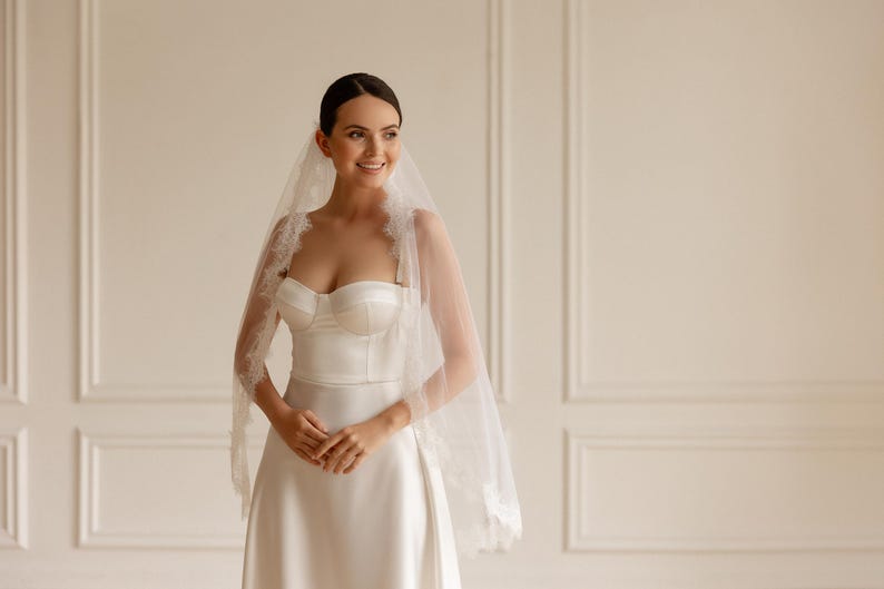 Puede incluir: Una novia con un vestido de novia de sat&eacute;n blanco y velo. El corpi&ntilde;o sin tirantes y la falda en forma de A crean una silueta elegante. El velo est&aacute; adornado con delicado encaje, a&ntilde;adiendo un toque rom&aacute;ntico.