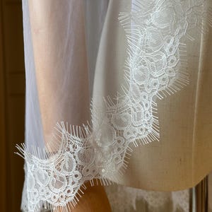 Puede incluir: Primer plano de una tela blanca transparente con un delicado ribete de encaje. El encaje presenta intrincados patrones florales y un borde festoneado. La tela est&aacute; drapeada sobre un maniqu&iacute;, mostrando la textura y el dise&ntilde;o.
