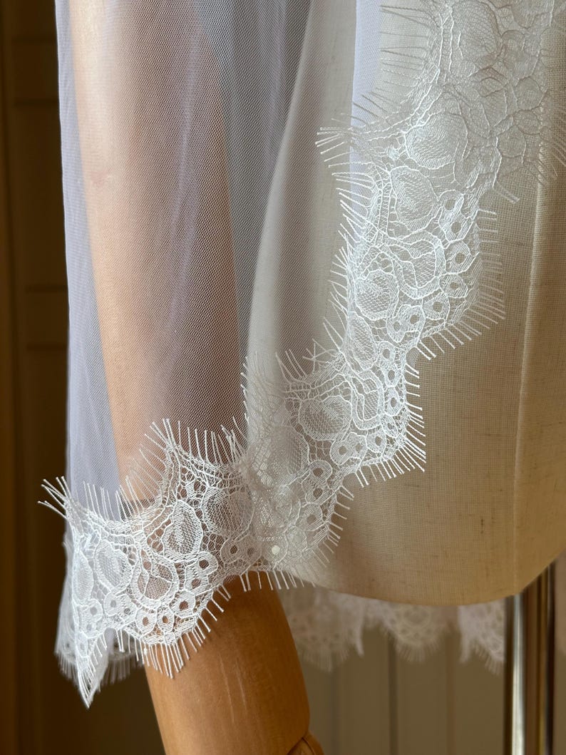 Puede incluir: Primer plano de una tela blanca transparente con un delicado ribete de encaje. El encaje presenta intrincados patrones florales y un borde festoneado. La tela est&aacute; drapeada sobre un maniqu&iacute;, mostrando la textura y el dise&ntilde;o.