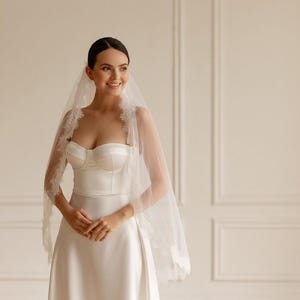 K&ouml;nnte beinhalten: Eine Braut in einem wei&szlig;en Brautkleid aus Satin mit einem herzf&ouml;rmigen Ausschnitt und einem passenden Schleier mit Spitzenbesatz. Der Schleier ist &uuml;ber die Schultern drapiert. Der Hintergrund ist eine hellbeige Wand.
