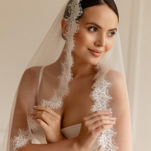 May include: A sheer white bridal veil with delicate lace trim. The veil is draped over the head and shoulders, with the lace detail framing the face. The bride is wearing a white dress.