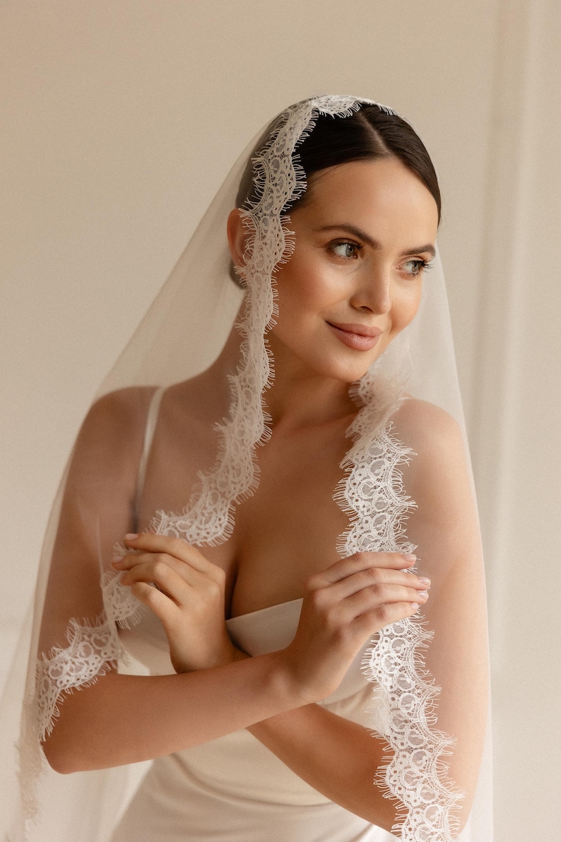 May include: A sheer white bridal veil with delicate lace trim. The veil is draped over the head and shoulders, with the lace detail framing the face. The bride is wearing a white dress.