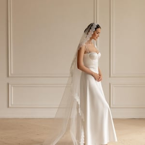 May include: A white bridal gown with a sweetheart neckline and spaghetti straps. The dress is paired with a long, sheer veil trimmed with delicate lace. The bride is wearing white heels. The background is a neutral color.