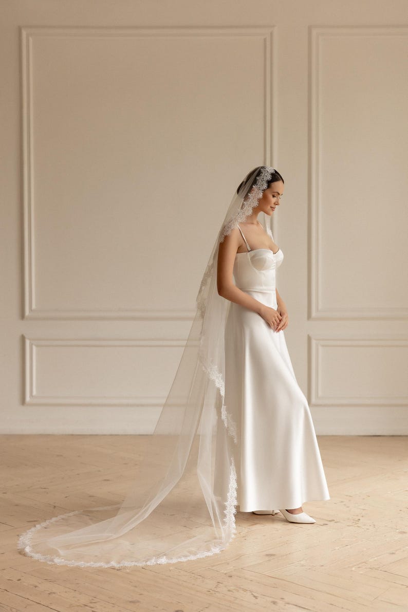 May include: A white bridal gown with a sweetheart neckline and spaghetti straps. The dress is paired with a long, sheer veil trimmed with delicate lace. The bride is wearing white heels. The background is a neutral color.