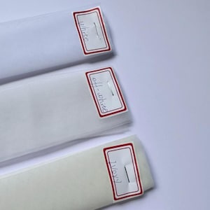 Puede incluir: Tres rollos de tela en blanco, blanco roto y marfil. Cada rollo tiene una etiqueta con el nombre del color. La tela blanca est&aacute; en la parte superior, seguida del blanco roto y el marfil en la parte inferior. La tela est&aacute; enrollada.