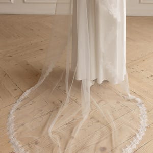May include: A long, sheer white wedding veil with a delicate lace trim. The veil is draped over a white dress, creating a flowing, elegant look. The floor is made of wooden planks.